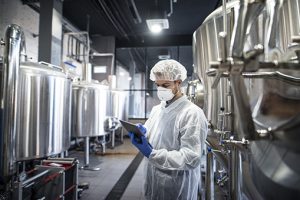 A man wearing protective clothing and a protective mask stands between large metal containers in a hall