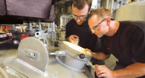 Two men with goggles look into a potion through a round glass pane on the tank